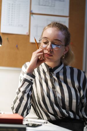 Photo of thinking young woman architect wearing glasses designing draft and sitting at workplace in...