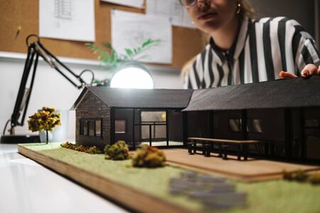 Photo of thoughtful caucasian woman architect wearing glasses designing draft with house model and...