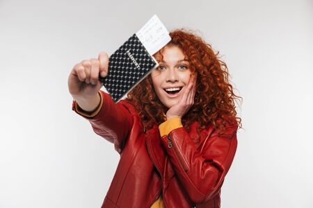 Portrait of elegant redhead woman 20s wearing leather jacket holding passport and travel tickets isolated over white backgroundの写真素材
