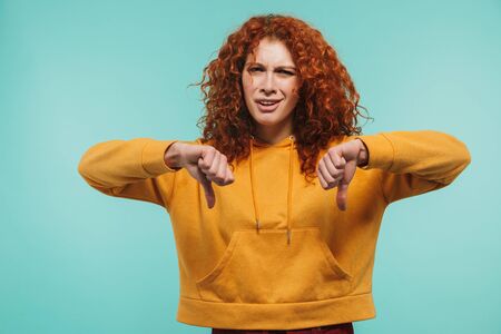Portrait of confused redhead woman 20s showing thumbs down and expressing dislike isolated over blue backgroundの写真素材
