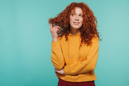 Image of gorgeous woman 20s with curly ginger hair smiling and looking at camera isolated over blue...