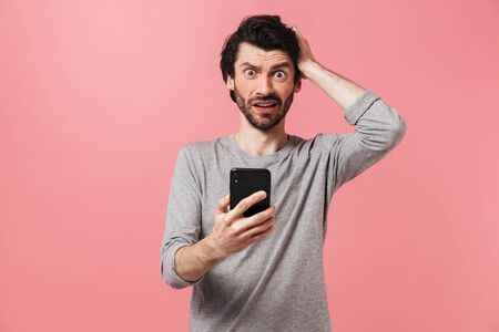 Image of a handsome confused serious young man isolated over pink wall background using mobile...
