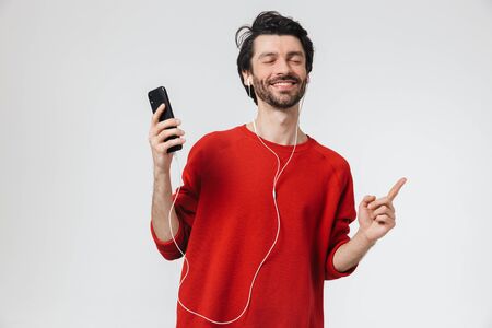Image of a handsome young excited man posing isolated over white wall background listening music with earphones using mobile phone.の写真素材