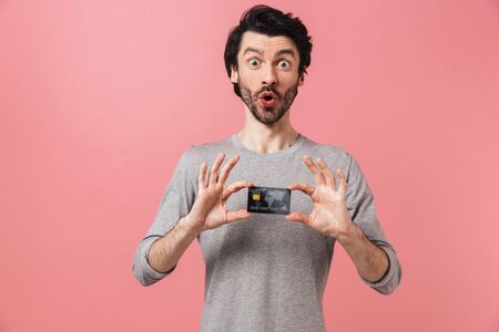 Handsome excited young bearded brunette man wearing sweater standing isolated over pink background, showing credit cardの写真素材