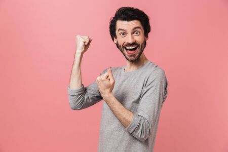 Handsome shocked young bearded brunette man wearing sweater standing isolated over pink background, celebratingの写真素材
