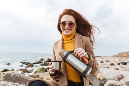 Lovely young redhead woman wearing autumn coat walking at the beach, drinking hot coffee from vaccum flaskの写真素材