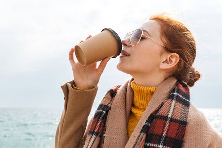 Beautiful young redheaded woman wearing coat walking at the beach, drinking takeaway coffee