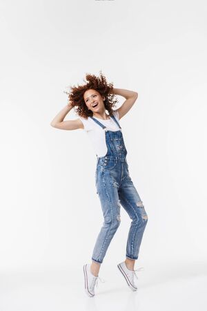 Full length of a cheerful young redhead curly haired girl wearing denim overalls posing isolated over white backgroundの写真素材