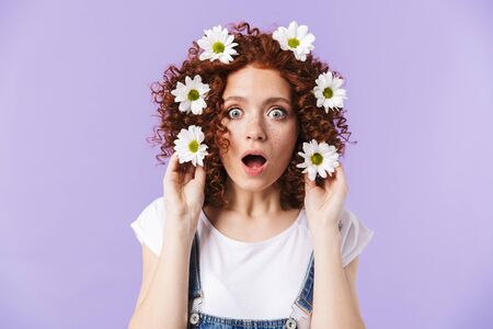 Image of a beautiful curly shocked redhead girl posing isolated over purple background with flowers in hair.の写真素材
