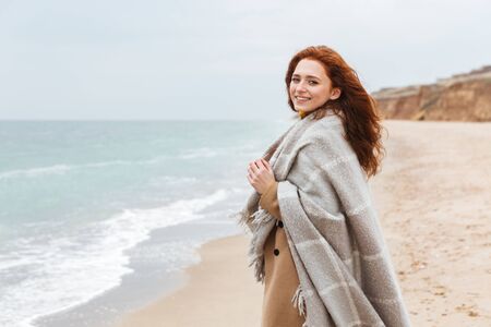 Beautiful young redheaded woman wearing coat, covered in blanket walking at the beach