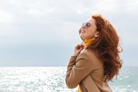 Beautiful young redheaded woman wearing coat walking at the beachの写真素材