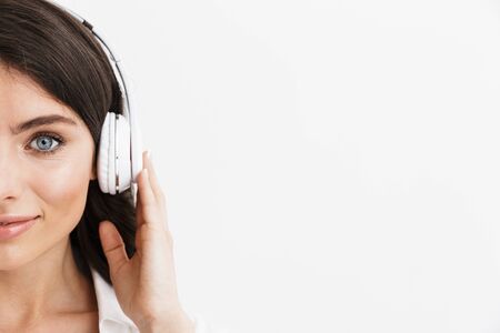 Close up half face of a beautiful young woman with long curly brunette hair wearing white shirt standing isolated over white background, enjoying listening to music with headphonesの写真素材
