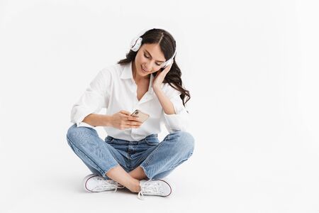 Cheerful young brunette woman wearing shirt sitting isolated over white background, listening to music with earphones and mobile phoneの写真素材