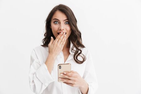 Beautiful surprised young woman with long curly brunette hair wearing white shirt standing isolated over white background, using mobile phoneの写真素材
