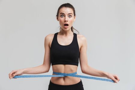 Image of a beautiful young sport fitness shocked woman posing with centimeter over waist isolated...