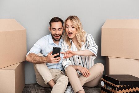 Photo of excited couple in casual clothing seating near cardboard boxes and using cellphone isolated...