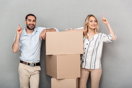 Photo of laughing couple in casual clothing standing near cardboard boxes and pointing fingers...