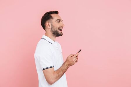 Side view of portrait of a happy handsome man standing isolated over pink background, holding mobile...
