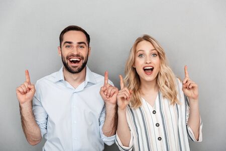 Photo closeup of excited couple in casual clothing smiling while pointing fingers upward at copyspace isolated over gray backgroundの写真素材