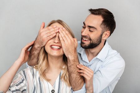 Photo closeup of handsome man in casual clothing smiling and covering eyes of his girlfriend isolated over gray wallの写真素材