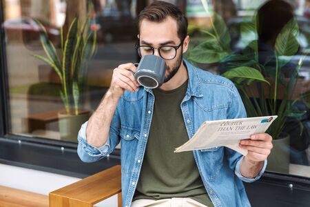 Photo of focused brunette man wearing denim shirt and glasses reading newspaper with cup of coffee in cafe outdoorsの写真素材