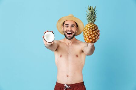 Photo of young shirtless tourist man wearing straw hat smiling while holding exotic fruits isolated...