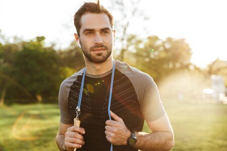 Image of a handsome young strong sports man posing outdoors at the nature park location with skipping rope.の写真素材