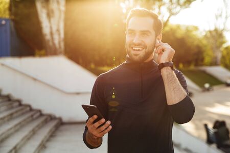 Image of a handsome happy young strong sports man posing outdoors at the nature park location using mobile phone listening music with earphones.の写真素材