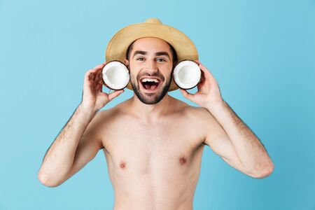 Photo of excited shirtless tourist man wearing straw hat smiling while holding two coconut parts isolated over blue backgroundの写真素材