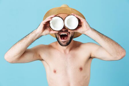 Photo of european shirtless tourist man wearing straw hat smiling while holding two coconut parts isolated over blue backgroundの写真素材