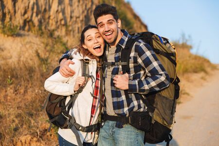 Cheerful young couple carrying backpacks hiking together, walking on a trail, huggingの写真素材