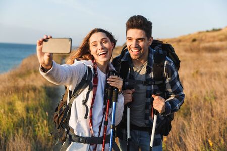Cheerful young couple carrying backpacks hiking together, walking on a trail, taking a selfieの写真素材