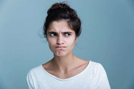 Close up portrait of a pensive unsatisfied pretty young brunette woman wearing white t-shirt standing isolated over blue backgroundの写真素材