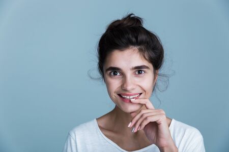Close up portrait of a pretty young brunette woman wearing white t-shirt standing isolated over blue background, posing, touching faceの写真素材