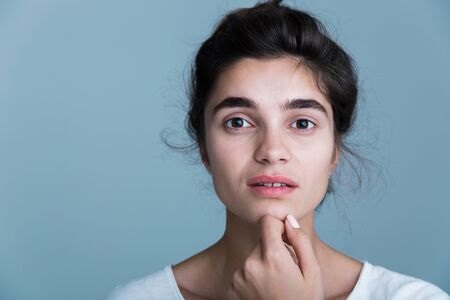 Close up portrait of a pretty young brunette woman wearing white t-shirt standing isolated over blue background, posing, touching faceの写真素材