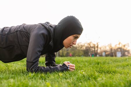 Image of a young amazing serious muslim sports fitness woman dressed in hijab and dark clothes outdoors in green nature park make exercises plank.の写真素材