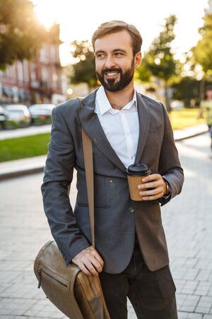 Portrait of caucasian adult businessman in formal suit holding takeaway coffee while walking through city streetの写真素材