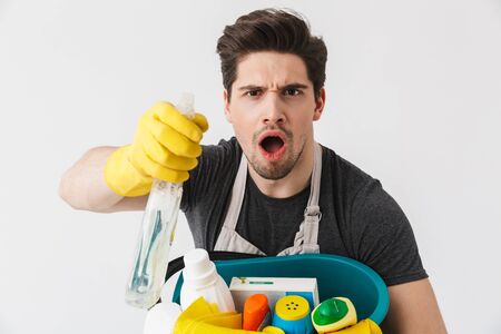 Handsome brunette houseman wearing apron standing isolated over white background, holding bucket full of detergentsの写真素材