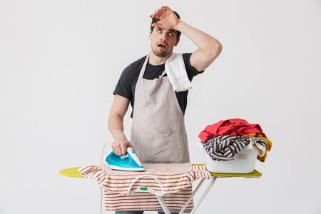 Handsome tired brunette houseman wearing apron standing isolated over white background, ironing clothes on a boardの写真素材