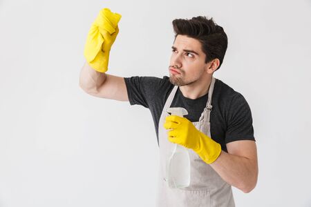 Handsome brunette houseman wearing apron standing isolated over white background, cleaning with a spongeの写真素材