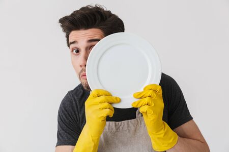 Photo of caucasian young man wearing yellow rubber gloves for hands protection washing dishes while cleaning house isolated over white backgroundの写真素材