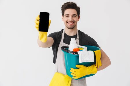 Handsome brunette houseman wearing apron standing isolated over white background, holding bucket full of detergents, showing blank screen mobile phoneの写真素材