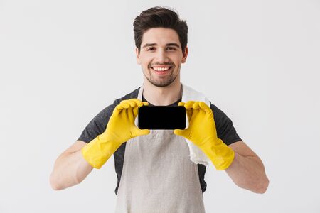 Handsome brunette houseman wearing apron standing isolated over white background, showing mobile phoneの写真素材