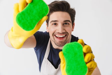 Handsome brunette houseman wearing apron standing isolated over white background, holding spongesの写真素材