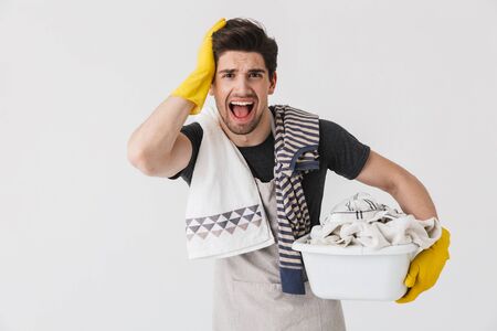 Photo of irritated young man wearing yellow rubber gloves doing laundry and carrying basket with dirty clothing isolated over white backgroundの写真素材