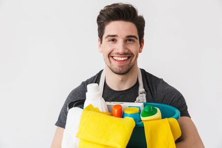 Handsome brunette houseman wearing apron standing isolated over white background, holding bucket full of detergentsの写真素材