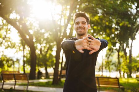 Image of a handsome cheerful young sports fitness man standing in green park nature make stretching exercises for arms.の写真素材