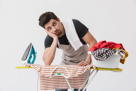 Image of a tired bored young houseman househusband with iron and clothes isolated over white wall background.の写真素材