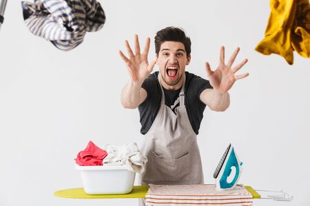 Handsome brunette houseman wearing apron standing isolated over white background, ironing clothes on a boardの写真素材