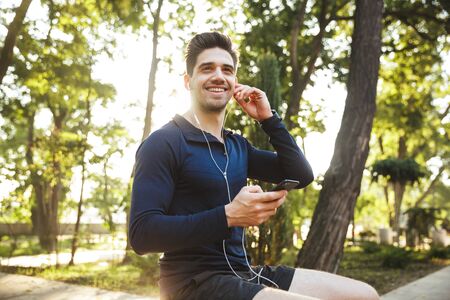 Portrait of attractive athletic man dressed in sportswear listening to music with earphones and smartphone while sitting on bench after workout in sunny green parkの写真素材